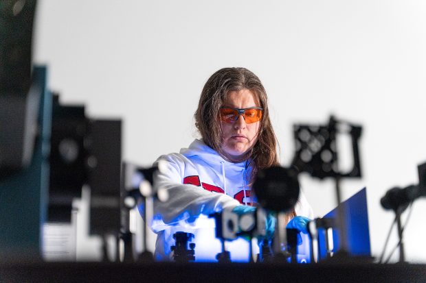 Woman with long brown hair wears orange-tinted glasses, a white SDSU sweatshirt and blue gloves while setting up a laser experiment. There is a blue glow emanating from the table she is standing behind. (Photo credit: Scott Hargrove)