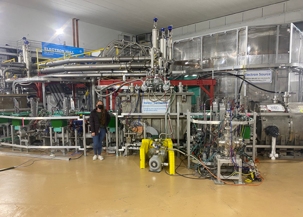 Dayna stands in front of a linear accelerator which is a large machine with several pipes and wires in a large room. She has long hair and is wearing a face mask, puffy dark jacket and jeans.