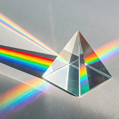 a clear glass equalateral triangle prism defracting a beam of white light into red, orange, yellow, green, indigo, and violet rays; on a gray background.